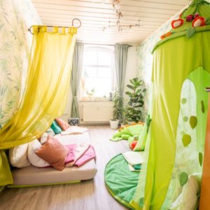 Heller Raum mit grünen Vorhängen und Baldachin, darunter Matratze mit vielen Kissen und Plüschtieren